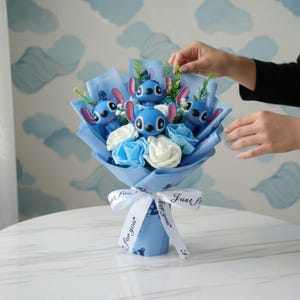 Pu&ograve; includere: Un bouquet con personaggi blu di Stitch e rose blu e bianche, avvolto in carta blu e legato con un nastro bianco con la scritta "Just for you". Il bouquet &egrave; su un tavolo bianco.