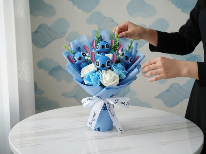 Pu&ograve; includere: Un bouquet con personaggi blu di Stitch e rose blu e bianche, avvolto in carta blu e legato con un nastro bianco con la scritta "Just for you". Il bouquet &egrave; su un tavolo bianco.