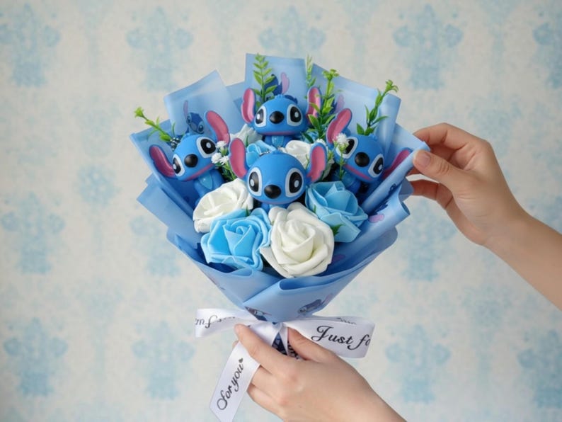 Pu&ograve; includere: Un bouquet con statuette blu di Stitch, rose di stoffa bianche e blu e verde, avvolto in carta blu. Un nastro bianco con la scritta "Just for you" &egrave; legato intorno alla base.