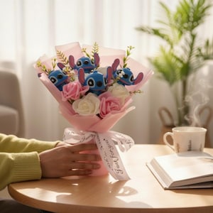 May include: A bouquet featuring blue character plushies, pink and white roses, and greenery, wrapped in pink paper and tied with a ribbon that reads "Just for you". The bouquet is on a wooden table.