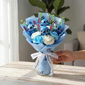 Pu&ograve; includere: Un bouquet con personaggi blu dei cartoni animati, carta blu e bianca e rose bianche e blu. Il bouquet &egrave; avvolto con un nastro blu con la scritta "For you". Il bouquet &egrave; in un vaso di vetro trasparente.