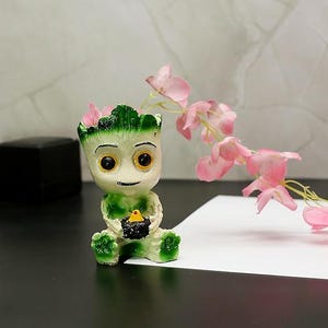 May include: A figurine of a cartoon character with a light green and cream color scheme, holding a small black object. The figurine has large yellow eyes and is adorned with pink flowers. A sprig of pink flowers is to the right.