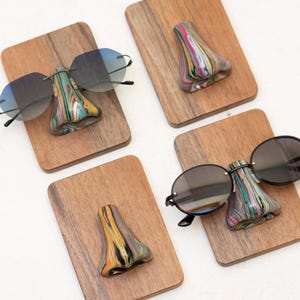 Puede incluir: Cuatro tableros de exhibición de madera, cada uno con un colorido soporte para gafas con forma de nariz. Dos tableros sostienen gafas de sol con lentes oscuras. Los soportes están pintados con una mezcla de colores, incluyendo rosa, amarillo, verde y negro.