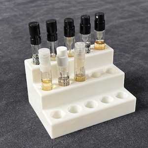 May include: A white tiered plastic display stand holding eight small glass perfume sample vials. The vials feature black or white spray caps and contain various shades of amber and clear liquid, arranged in two rows on the stepped structure.