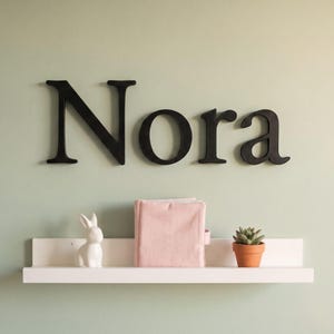 Op de afbeelding: Zwarte muurletters die de naam Nora vormen, geplaatst boven een witte zwevende plank. Op de plank staan een wit keramieken konijnenbeeldje, een zachtroze stoffen boek en een kleine groene vetplant in een terracotta pot.