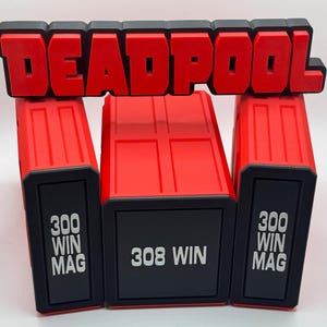 May include: A red and black Deadpool sign positioned above three rectangular storage containers. The two outer containers are labeled 300 WIN MAG in white text, while the center container is labeled 308 WIN in white text on a black background.
