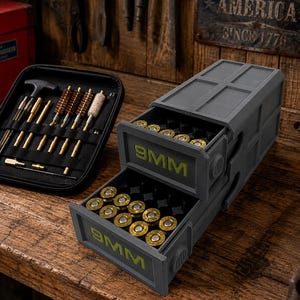 May include: A dark gray plastic storage container with two pull-out drawers, each labeled with green 9MM text, filled with brass-cased ammunition. Beside it sits a black zippered case containing various brass cleaning rods and brushes.