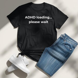 May include: Black t-shirt with the text "ADHD loading... please wait" in white, alongside blue jeans and white trainers. The t-shirt is knotted at the waist. The jeans are a light wash denim.