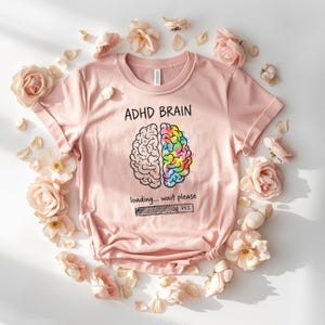 May include: A pale pink t-shirt featuring the text "ADHD BRAIN" and a brain graphic. The brain is split, with one half in black and white and the other in rainbow colours. Below it reads "loading... wait please" with a 75% loading bar.