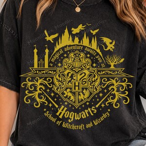 Può includere: Una t-shirt a maniche corte color nero antracite con una stampa grafica dorata dello stemma di Hogwarts, sagome di castelli e creature volanti. Il testo recita, a magical adventure awaits you at Hogwarts School of Witchcraft and Wizardry.