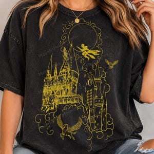 May include: A charcoal black short-sleeve t-shirt featuring a gold-coloured line art graphic of a large castle, a figure flying on a broomstick, a soaring owl, and a winged creature. The design includes swirling cloud motifs and a circular moon.