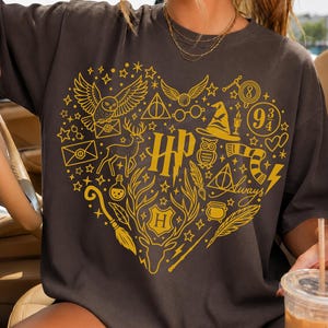 May include: A dark grey short-sleeved t-shirt featuring a large heart-shaped graphic in gold ink. The design includes various icons such as an owl, a stag, a sorting hat, a broomstick, a wand, the letters HP, and the number 9 3/4, all arranged in a heart.