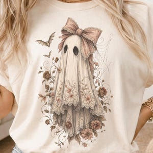 May include: A cream-colored t-shirt featuring a graphic print of a ghost draped in lace fabric with a large bow on its head. The ghost is surrounded by intricate illustrations of roses, vines, spiderwebs, and two small flying bats in muted tones.