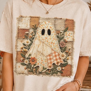May include: A cream-coloured short-sleeve t-shirt featuring a central square graphic of a ghost made from a patchwork of floral and checkered fabrics. The ghost is surrounded by embroidered flowers and foliage in muted earth tones.