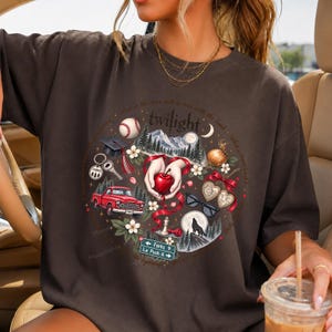 May include: A dark charcoal gray short-sleeve t-shirt featuring a circular graphic print with the word Twilight. The design includes a red truck, a baseball, a graduation cap, a red apple, sunglasses, a wolf silhouette, and a Forks road sign.