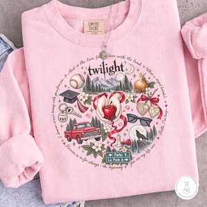May include: A light pink crewneck sweatshirt featuring a circular graphic print with the word twilight at the top. The design includes a red apple, a red truck, a baseball, a graduation cap, a wolf, sunglasses, and a road sign reading Forks 9 La Push 6.