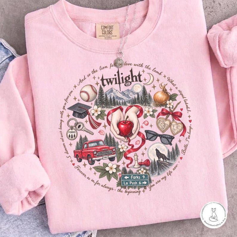 May include: A light pink crewneck sweatshirt featuring a circular graphic print with the word twilight at the top. The design includes a red apple, a red truck, a baseball, a graduation cap, a wolf, sunglasses, and a road sign reading Forks 9 La Push 6.