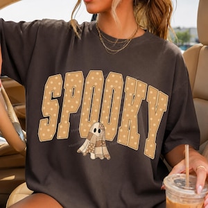 May include: A dark charcoal gray short-sleeve t-shirt featuring the word SPOOKY in large, tan, polka-dotted varsity-style letters. A small ghost character with a patchwork pattern of brown and tan fabric squares is positioned below the text.