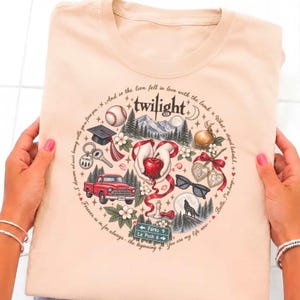 May include: A light peach-colored t-shirt featuring a circular graphic with the word twilight at the top. The design includes illustrations of a red apple, a red truck, a baseball, a graduation cap, sunglasses, a wolf, and a road sign reading Forks 9 La Push 6, surrounded by text quotes.