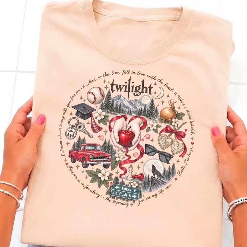 May include: A light peach-colored t-shirt featuring a circular graphic with the word twilight at the top. The design includes illustrations of a red apple, a red truck, a baseball, a graduation cap, sunglasses, a wolf, and a road sign reading Forks 9 La Push 6, surrounded by text quotes.