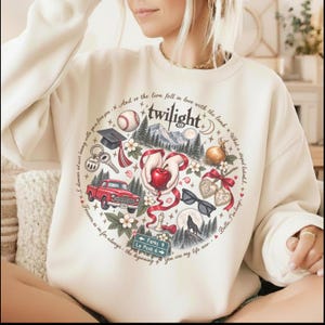 May include: A cream-colored crewneck sweatshirt featuring a circular graphic print. The design includes a red truck, a baseball, a graduation cap, a red apple held by hands, sunglasses, a wolf silhouette, and text reading Twilight and Forks.