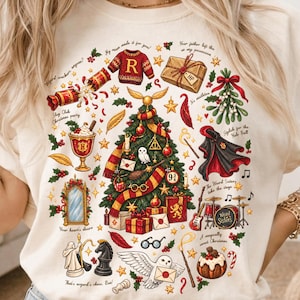 May include: A cream-coloured t-shirt featuring a festive graphic print of a Christmas tree decorated with wizarding-themed ornaments, including a golden snitch, a red and gold scarf, glasses, and various magical items, surrounded by holiday text.