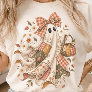 Könnte beinhalten: Ein cremefarbenes T-Shirt mit einer Grafik eines Geistes, der eine große orange-weiß karierte Schleife trägt. Der Geist hat einen Körper im Patchwork-Stil mit Blumen- und Karomustern in Orange, Grün und Gelb und trägt einen passenden Kürbiskorb.