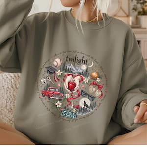 May include: An olive green crewneck sweatshirt featuring a circular graphic print. The design includes a red apple, a red truck, a baseball, a graduation cap, a wolf silhouette, sunglasses, and a sign reading Forks 9 La Push 6.