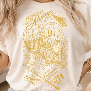 Pode incluir: Uma t-shirt cor de creme com uma impressão gráfica dourada de um castelo, um comboio a vapor, uma coruja e o texto Always, 9 3/4, Accio, Expelliarmus e Expecto patronum, juntamente com uma varinha e o símbolo dos Talismãs da Morte.