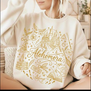 Peut inclure: Un sweat-shirt à col rond de couleur crème avec une impression graphique dorée. Le motif comprend un château, un hibou en vol, un balai, une pile de livres, une baguette et le mot Alohomora en écriture cursive, entouré de petites icônes.