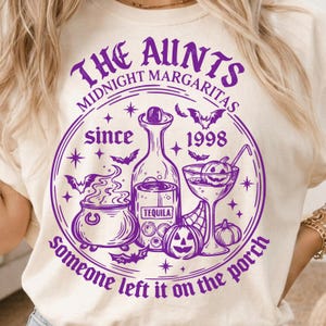 Puede incluir: Una camiseta color crema con un gráfico morado que incluye el texto THE AUNTS MIDNIGHT MARGARITAS since 1998 SOMEONE LEFT IT ON THE PORCH. El diseño muestra un caldero, una botella de tequila, una copa de margarita con una calabaza y murciélagos.