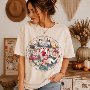 May include: A beige short-sleeve t-shirt featuring a circular graphic print with the word Twilight at the top. The design includes illustrations of a red truck, a baseball, a graduation cap, a red apple, sunglasses, a wolf, and road signs.