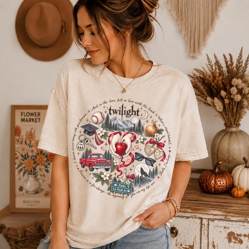 May include: A beige short-sleeve t-shirt featuring a circular graphic print with the word Twilight at the top. The design includes illustrations of a red truck, a baseball, a graduation cap, a red apple, sunglasses, a wolf, and road signs.