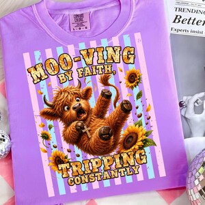 May include: A light purple t-shirt featuring a graphic of a brown Highland cow wearing a gold cross necklace. The cow is surrounded by sunflowers and vertical light blue stripes. The text reads MOO-VING BY FAITH, TRIPPING CONSTANTLY.
