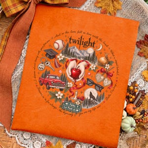 May include: An orange fabric item featuring a circular graphic with the text Twilight. The design includes a baseball, graduation cap, red truck, hands holding an apple, sunglasses, a wolf silhouette, and a road sign reading Forks 9 La Push 6.