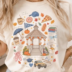 May include: A cream-colored t-shirt featuring a central gazebo illustration surrounded by various icons including a coffee mug, pizza slice, hot dog, Yale pennant, books, dragonfly, ballet slippers, graduation cap, and a yellow jeep with text.