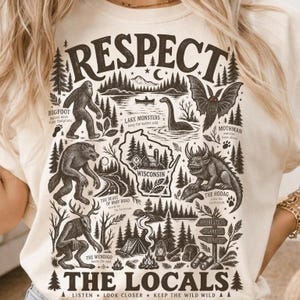 Op de afbeelding: Een crèmekleurig t-shirt met een donkerbruine grafische print van de tekst RESPECT THE LOCALS. Het ontwerp bevat illustraties van Bigfoot, een meermonster, Mothman, een weerwolf, de Hodag en de Wendigo rondom een kaart van Wisconsin.