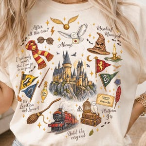 Op de afbeelding: Een crèmekleurig t-shirt met een gedetailleerde grafische print van een kasteel, een stoomtrein, een sorteerhoed, een bezemsteel, een uil en diverse magische voorwerpen. De tekst bevat zinnen zoals After all this time en Mischief Managed.