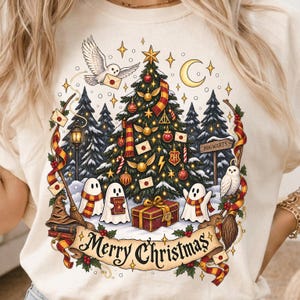 May include: A cream-coloured t-shirt featuring a festive graphic of a Christmas tree decorated with golden snitches, scarves, and letters. The design includes white owls, ghosts wearing scarves, a gift box, and a banner reading Merry Christmas.