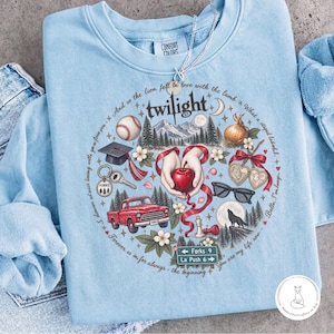 May include: A light blue crewneck sweatshirt featuring a circular graphic print with the word twilight at the top. The design includes a baseball, graduation cap, red truck, sunglasses, a wolf, and hands holding a red apple, surrounded by text.