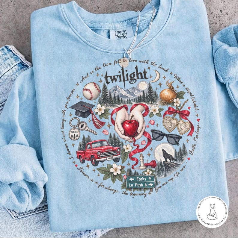 May include: A light blue crewneck sweatshirt featuring a circular graphic print with the word twilight at the top. The design includes a baseball, graduation cap, red truck, sunglasses, a wolf, and hands holding a red apple, surrounded by text.