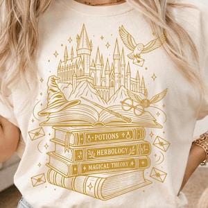 May include: A cream-coloured t-shirt featuring a gold-toned line art graphic of a castle, an owl carrying a letter, a wizard hat, a wand, round glasses, and a stack of books labelled Potions, Herbology, and Magical Theory with flying envelopes.