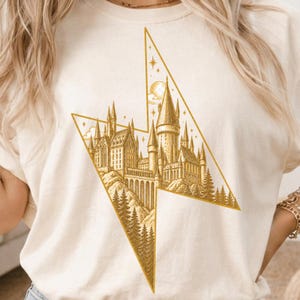 May include: A cream-coloured t-shirt featuring a gold-toned graphic print of a large castle with multiple towers and spires, set within a lightning bolt shape. The design includes detailed illustrations of surrounding pine trees and stars.
