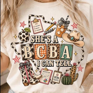 Op de afbeelding: Een crèmekleurig t-shirt met een grafische print met de tekst 'SHE'S A BCBA I CAN TELL'. Het ontwerp bevat een klembord met 'ABC' en 'Antecedent Behavior Consequence', een cowboyhoed, laarzen, een cactus, een drankje en potloden.