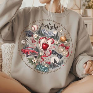 May include: A beige crewneck sweatshirt featuring a circular graphic print with the word twilight at the top. The design includes a red apple, a red truck, a baseball, a graduation cap, a wolf, sunglasses, and a road sign reading Forks 9 La Push 6.