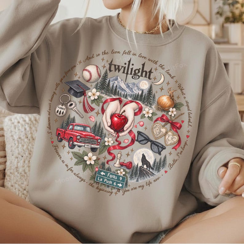 May include: A beige crewneck sweatshirt featuring a circular graphic print with the word twilight at the top. The design includes a red apple, a red truck, a baseball, a graduation cap, a wolf, sunglasses, and a road sign reading Forks 9 La Push 6.