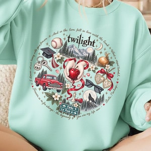 May include: A light mint green sweatshirt featuring a circular graphic with the text Twilight. The design includes a baseball, graduation cap, red truck, apple, sunglasses, wolf, and a street sign reading Forks 9 La Push 6, surrounded by forest imagery.