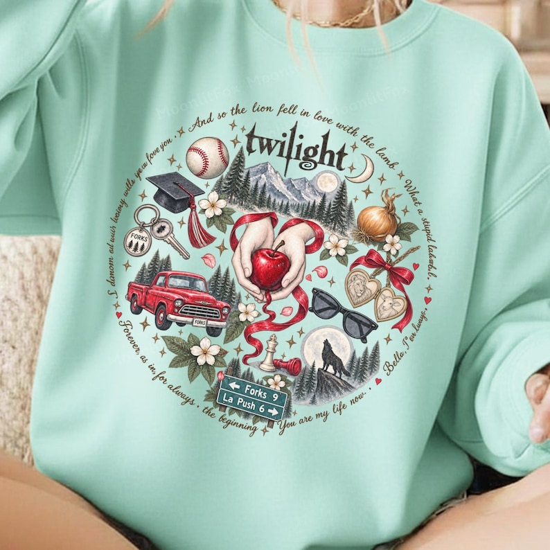 May include: A light mint green sweatshirt featuring a circular graphic with the text Twilight. The design includes a baseball, graduation cap, red truck, apple, sunglasses, wolf, and a street sign reading Forks 9 La Push 6, surrounded by forest imagery.
