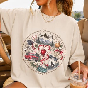 May include: A beige t-shirt featuring a circular graphic with the word twilight at the top. The design includes a baseball, graduation cap, red truck, apple, sunglasses, wolf, and a road sign reading Forks 9 and La Push 6, surrounded by text.