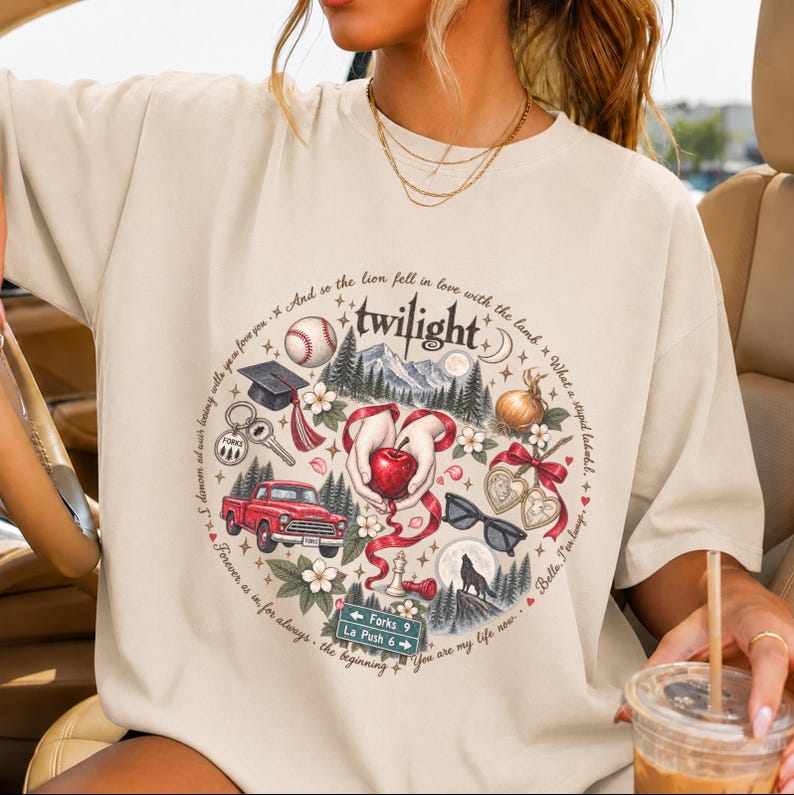 May include: A beige t-shirt featuring a circular graphic with the word twilight at the top. The design includes a baseball, graduation cap, red truck, apple, sunglasses, wolf, and a road sign reading Forks 9 and La Push 6, surrounded by text.